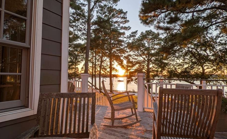 Wooden deck with chairs and a table overlooking a lake at sunset, surrounded by trees and a white railing.