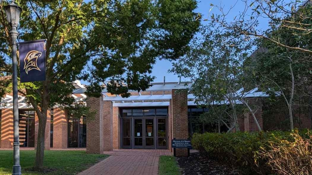A brick pathway leads to the entrance of a brick building with glass doors, surrounded by trees and shrubs. A pole with a bird logo banner stands on the left.