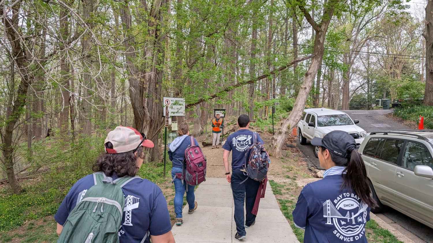 A group of people with backpacks walk on a paved path through a wooded area toward a person in an orange vest holding a sign.