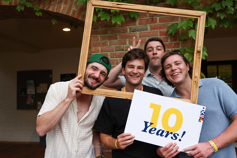 Four men smile for the camera, one holding a wooden frame and another holding a "10 Years!" sign, posing in front of a brick wall with green vines.