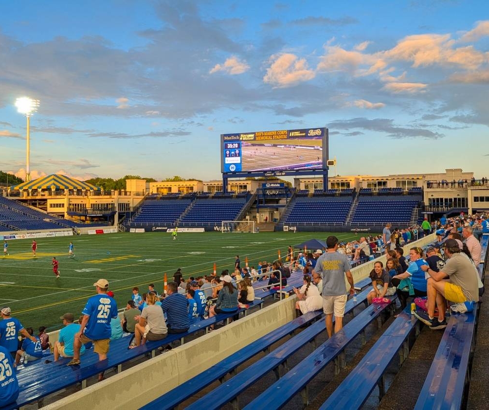 Fans sit in stadium bleachers watching a soccer match at sunset, with a large scoreboard and stadium lights visible in the background.
