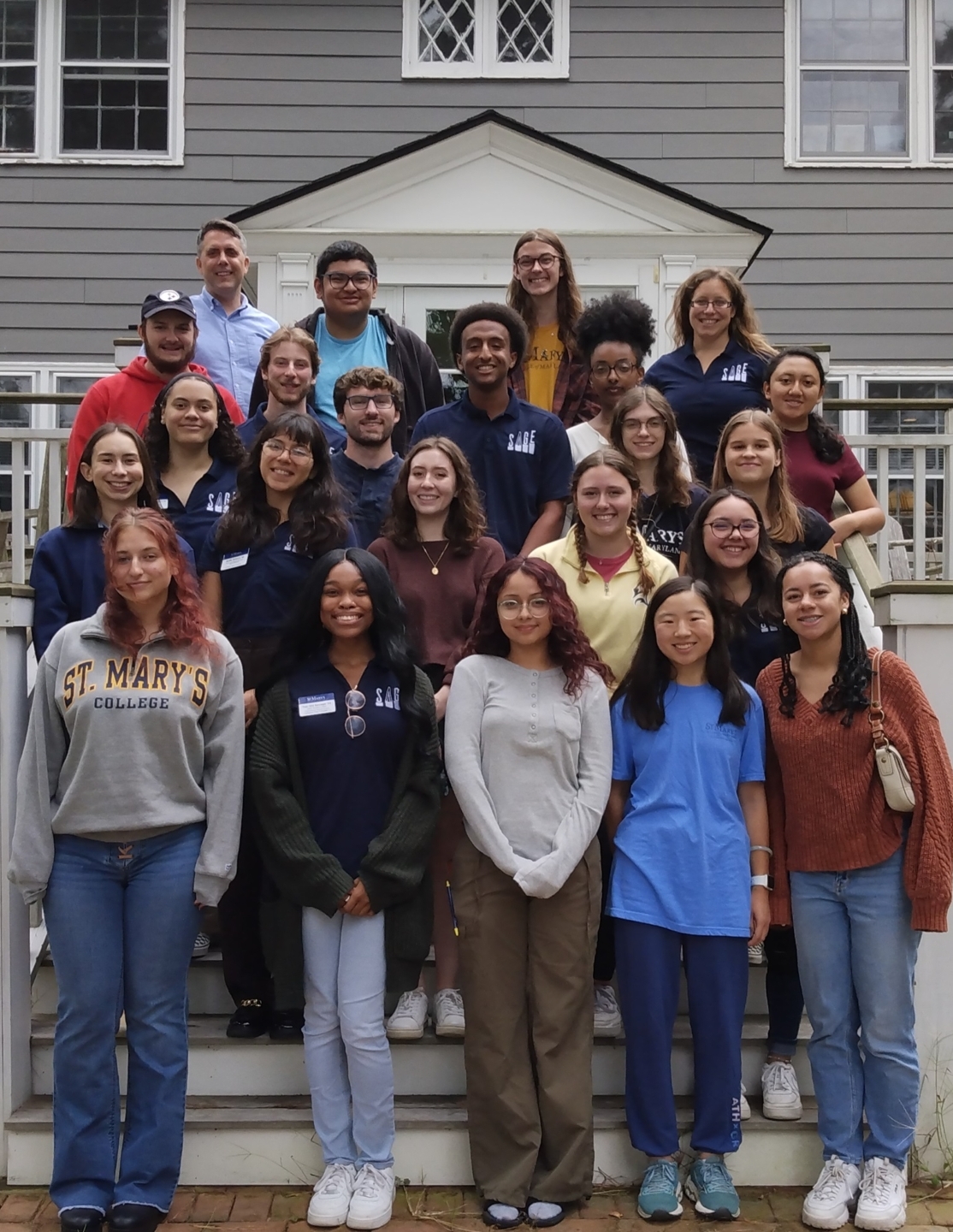 A group of 25 people pose together on the steps in front of a gray house with white trim and railings.