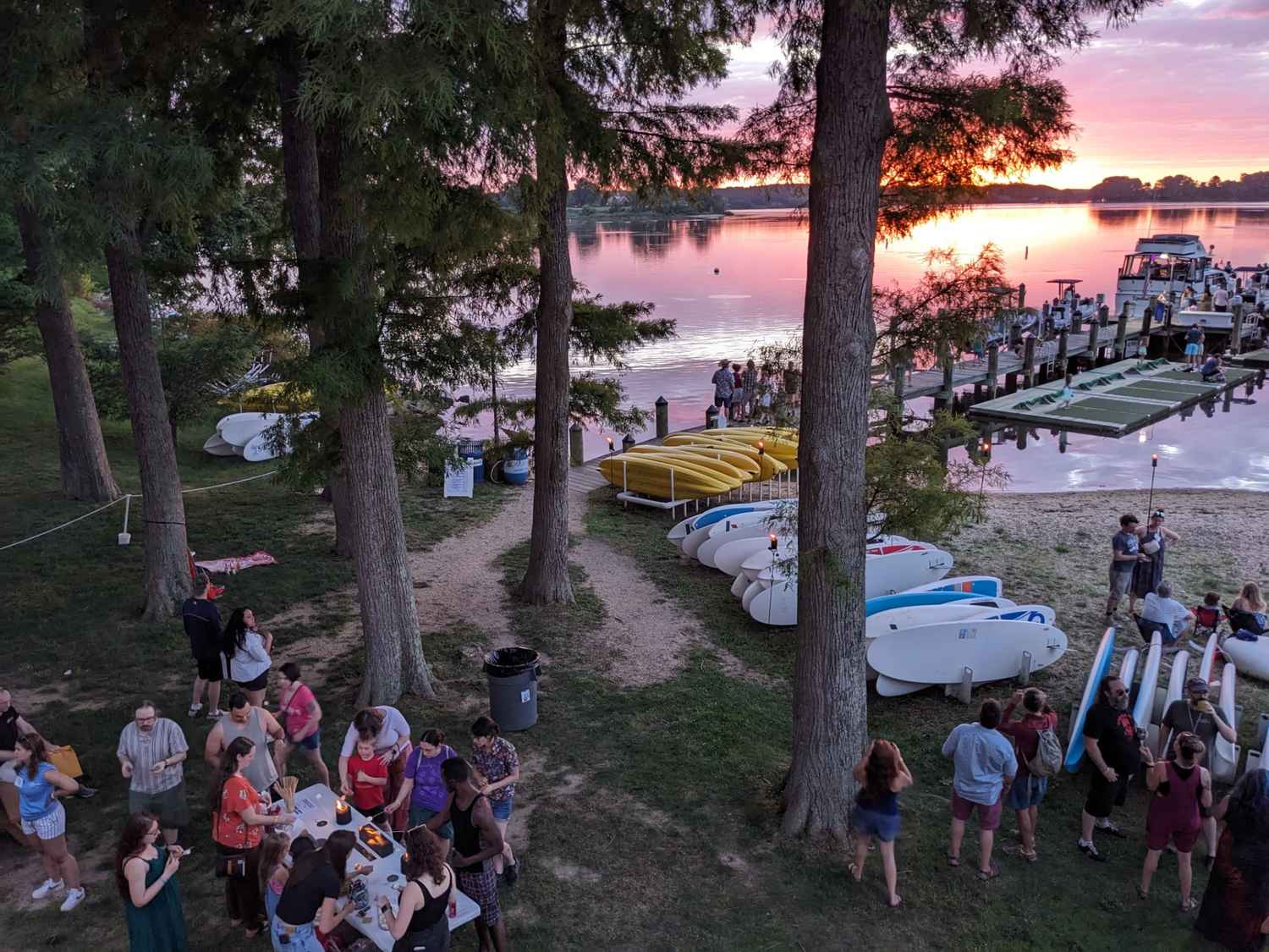 People gather near paddleboards and kayaks by a lakeshore at sunset, with groups socializing on the grass and boats docked in the water.