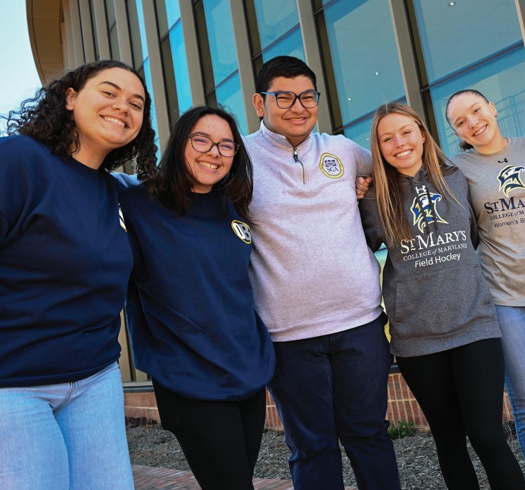 Six smiling students stand close together outside a building, wearing St. Mary’s College of Maryland athletic apparel.