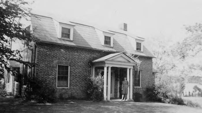 A black and white photo of the May Russell Alumni Lodge in 1941 when it was known as the Home EC Cottage, along with a student posing in the doorway