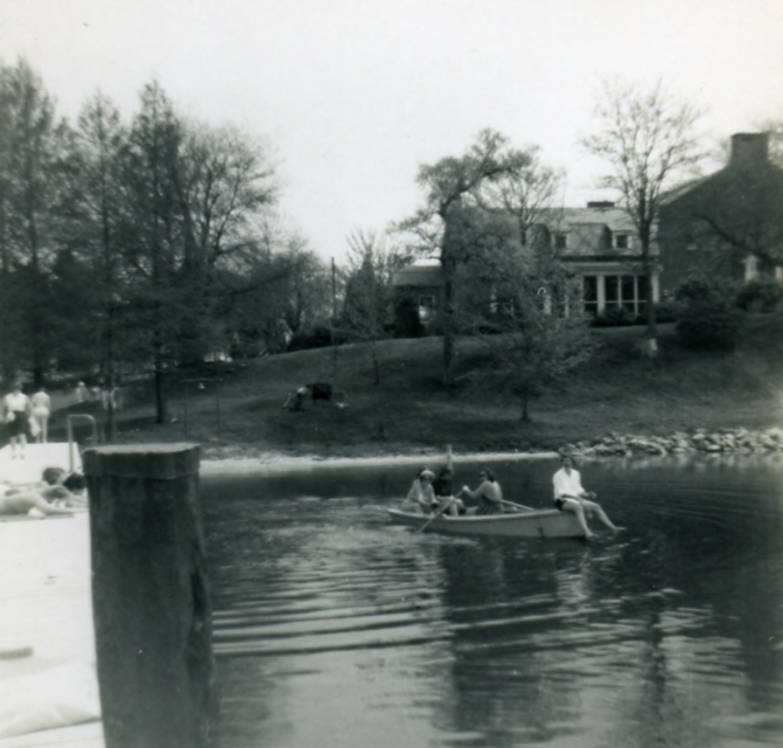 A black and white photo looking back at the May Russell Alumni Lodge from the waterfront dock in 1955, along with students on the docks, hillside, and in a rowboat in the river