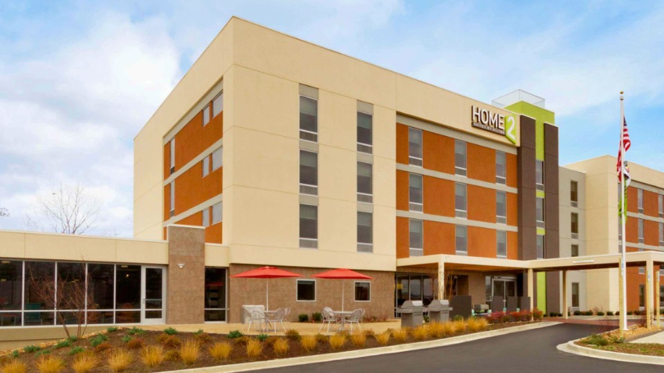A modern four-story hotel building with outdoor seating, red umbrellas, and an American flag, with the "Home2" sign visible on the facade.