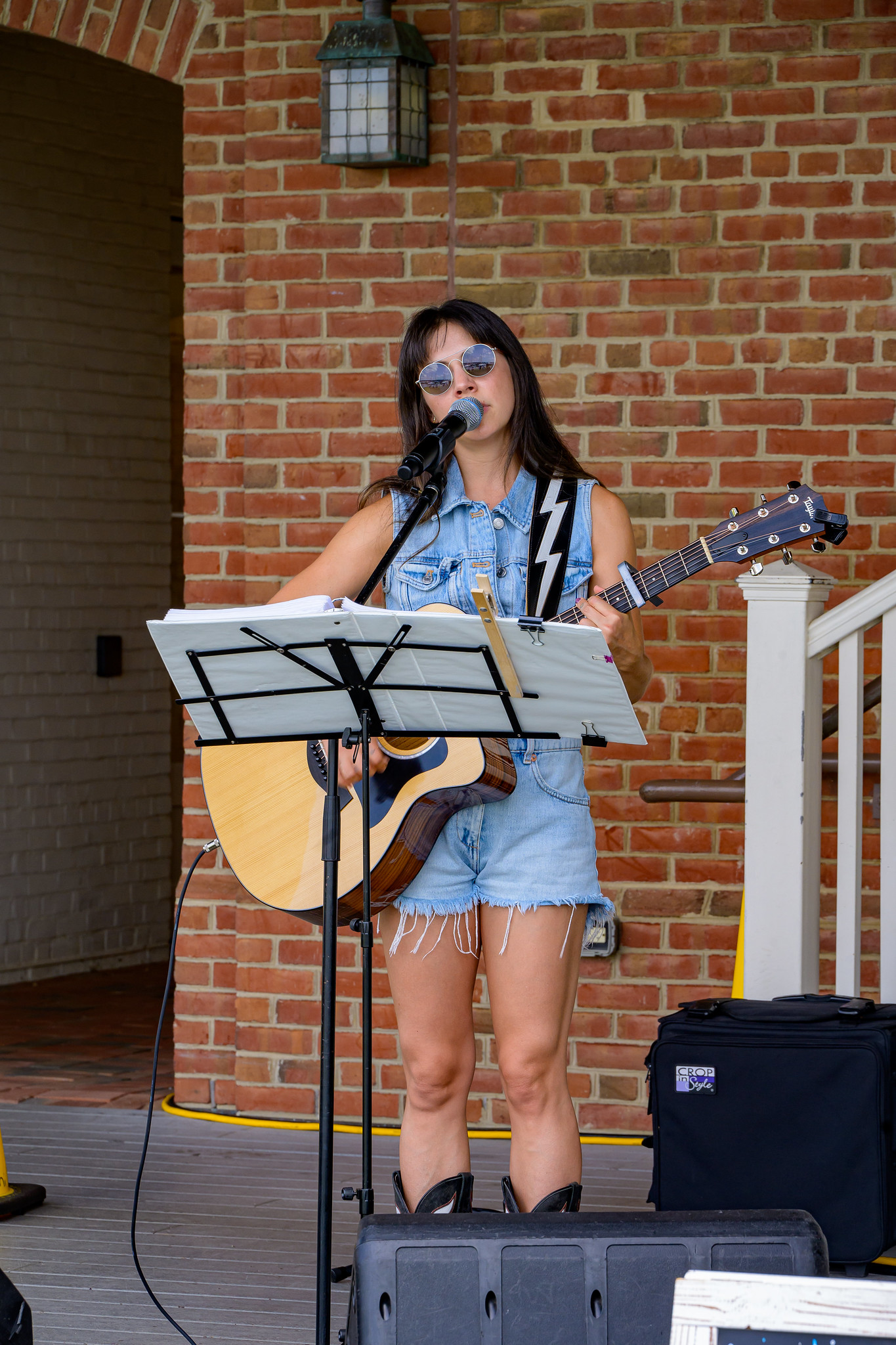 A woman wearing sunglasses and denim clothes sings and plays an acoustic guitar on an outdoor stage with a brick wall backdrop.