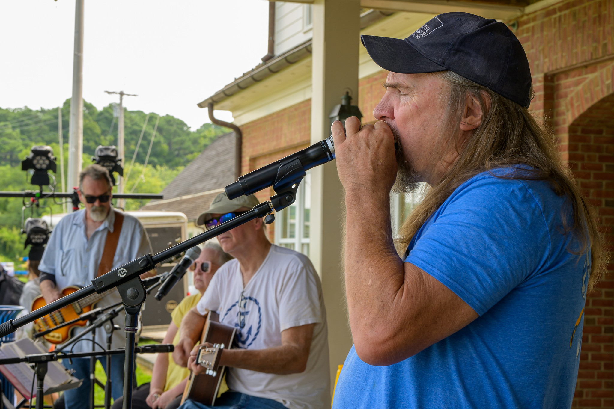 A man in a blue shirt and cap plays harmonica into a microphone while three other musicians perform on guitars outdoors.