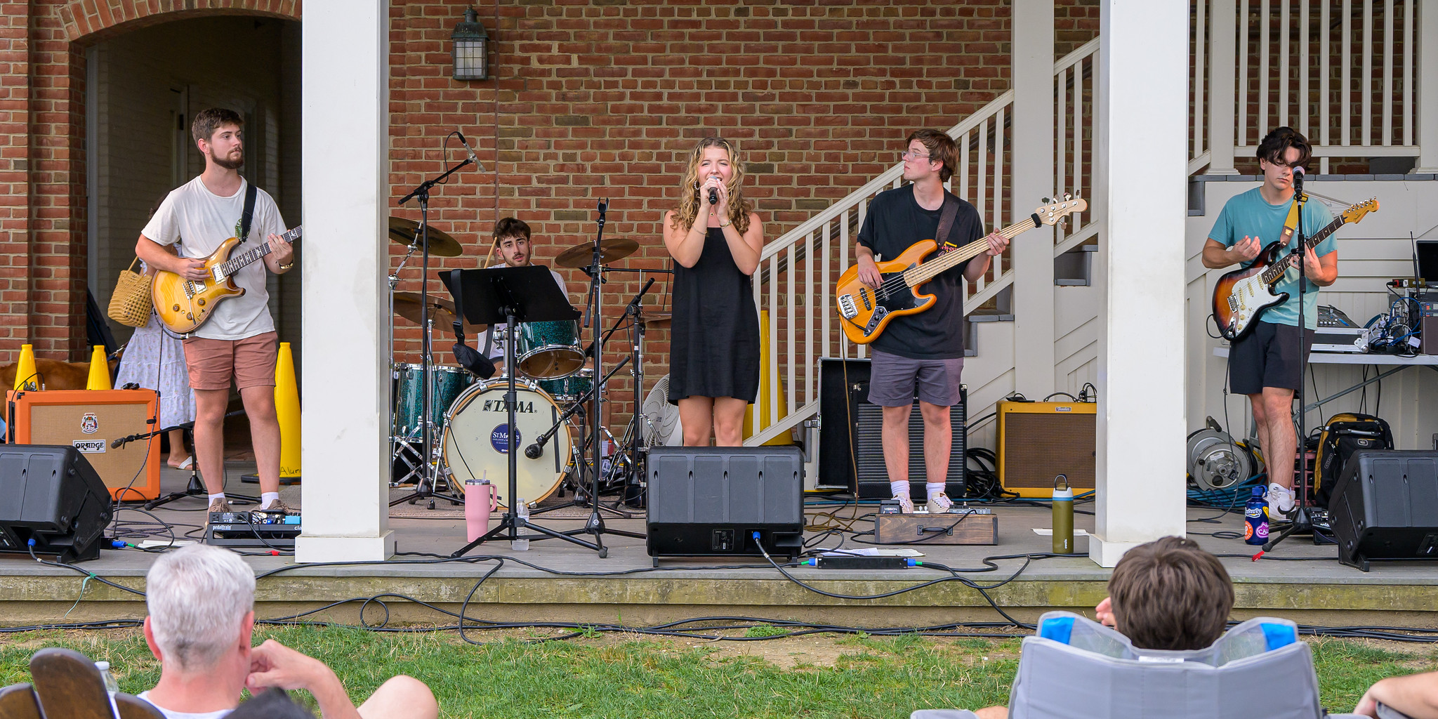A five-member band performs on an outdoor stage in front of a brick building while people watch from the grass.