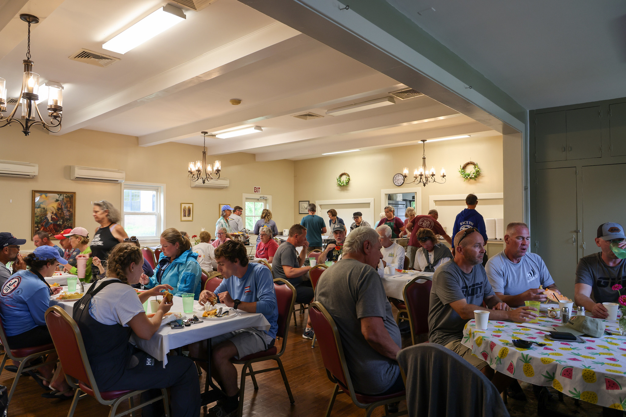 A group of people eat and socialize at tables in a brightly lit community dining hall decorated with floral tablecloths and wall art.