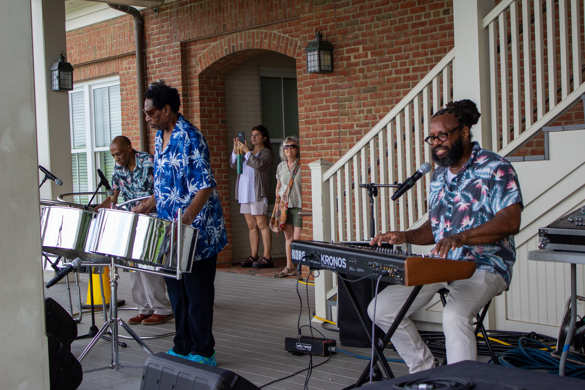 Three musicians perform on a covered patio, playing steel drums and keyboard, while a small audience stands nearby watching and taking photos.
