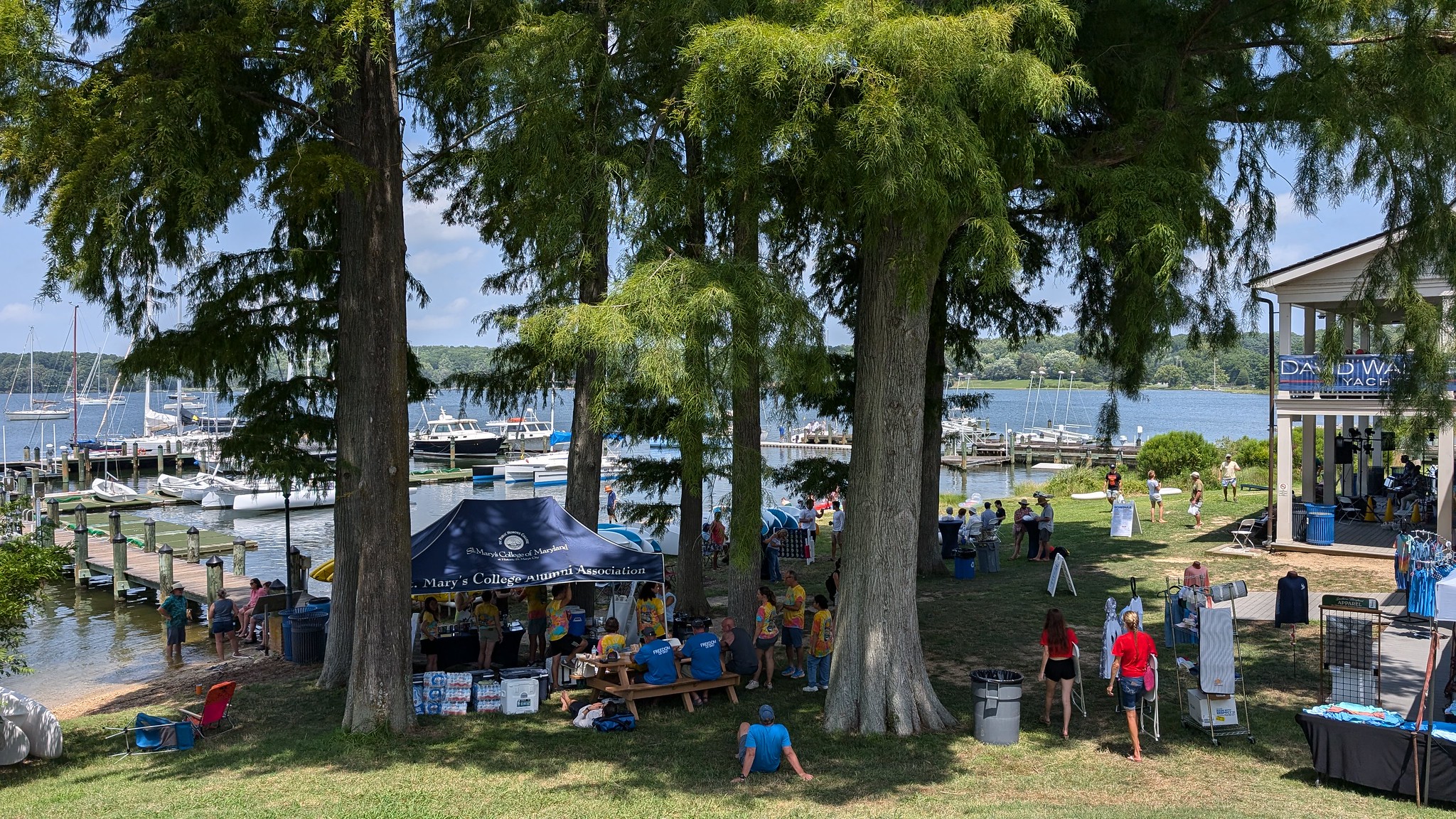 People gather under tall trees near a marina with boats docked, tents set up for an event, and a building with a "DAY ON THE WATER" sign.
