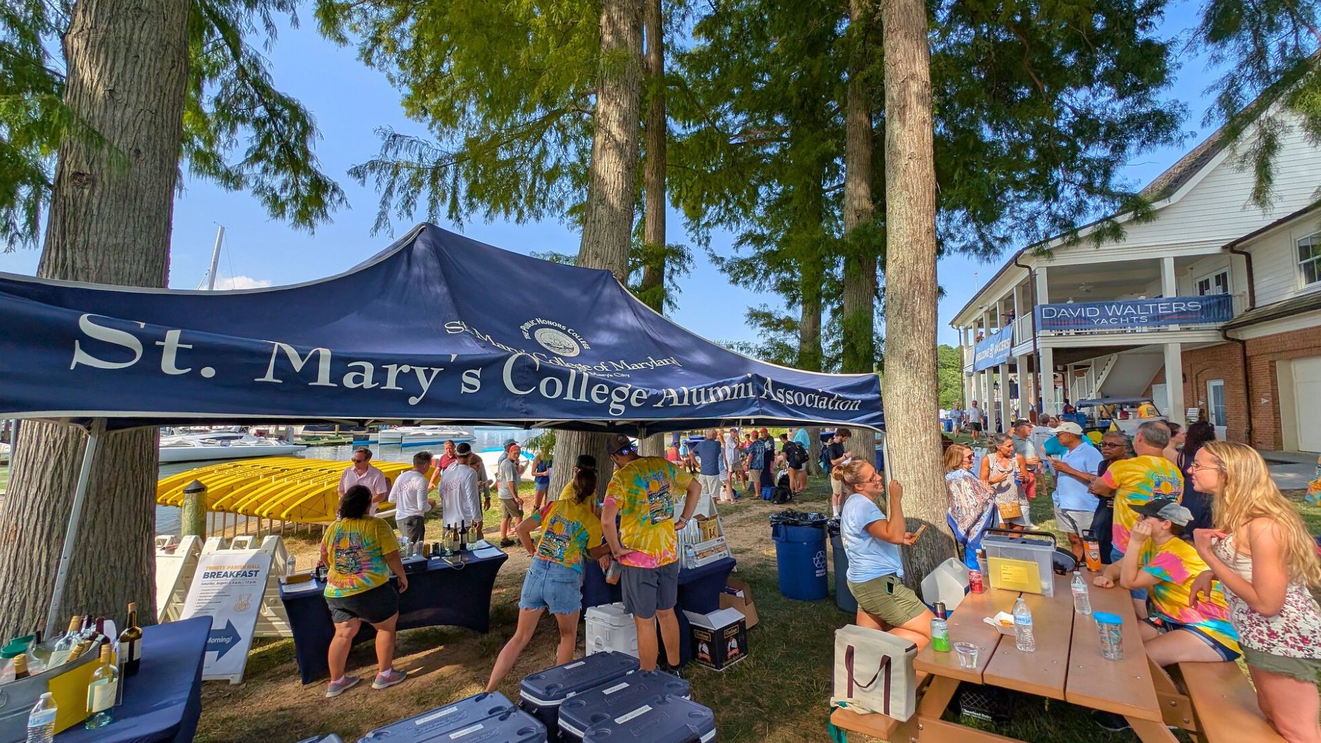 People gather under a St. Mary’s College Alumni Association tent at an outdoor event near a waterfront, with tables, coolers, and a building in the background.