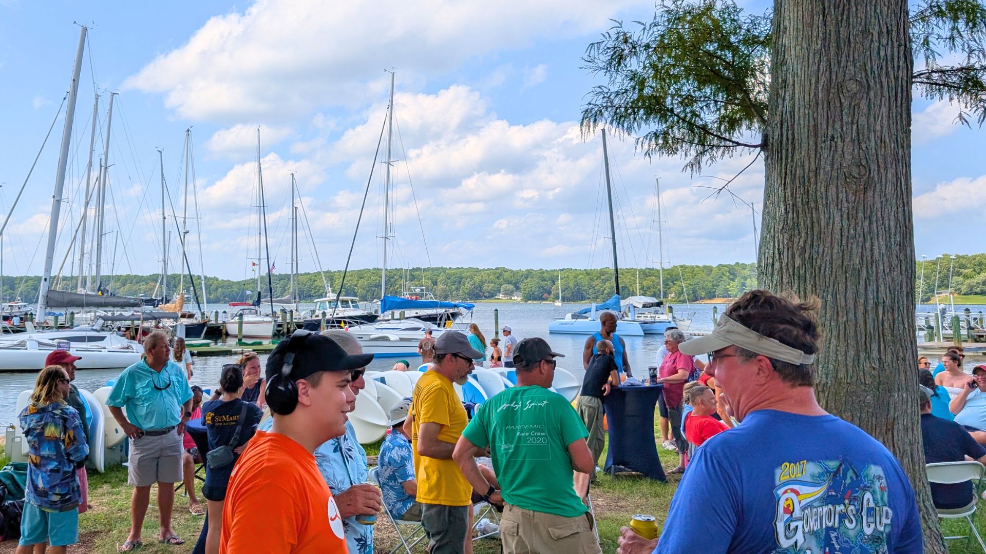 People gather by a marina with sailboats, talking and standing near trees on a sunny day. Some wear colorful event shirts and hats, with boats and water in the background.