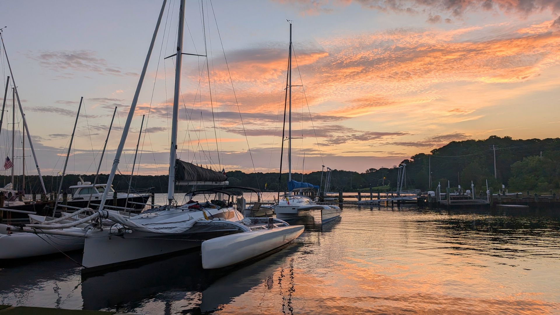 Catamarans and sailboats docked at a marina during sunset, with calm water reflecting the orange and pink hues of the sky.