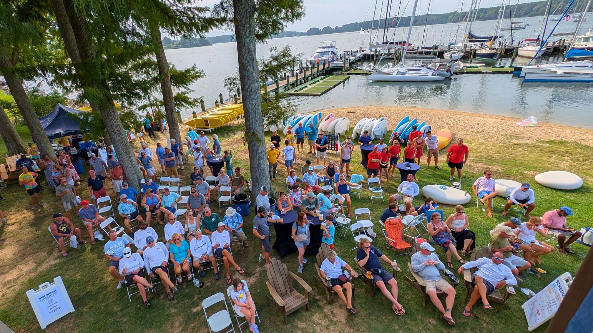 A group of people sits and stands outdoors near a marina with sailboats, kayaks, and canoes, attending an event by the water.