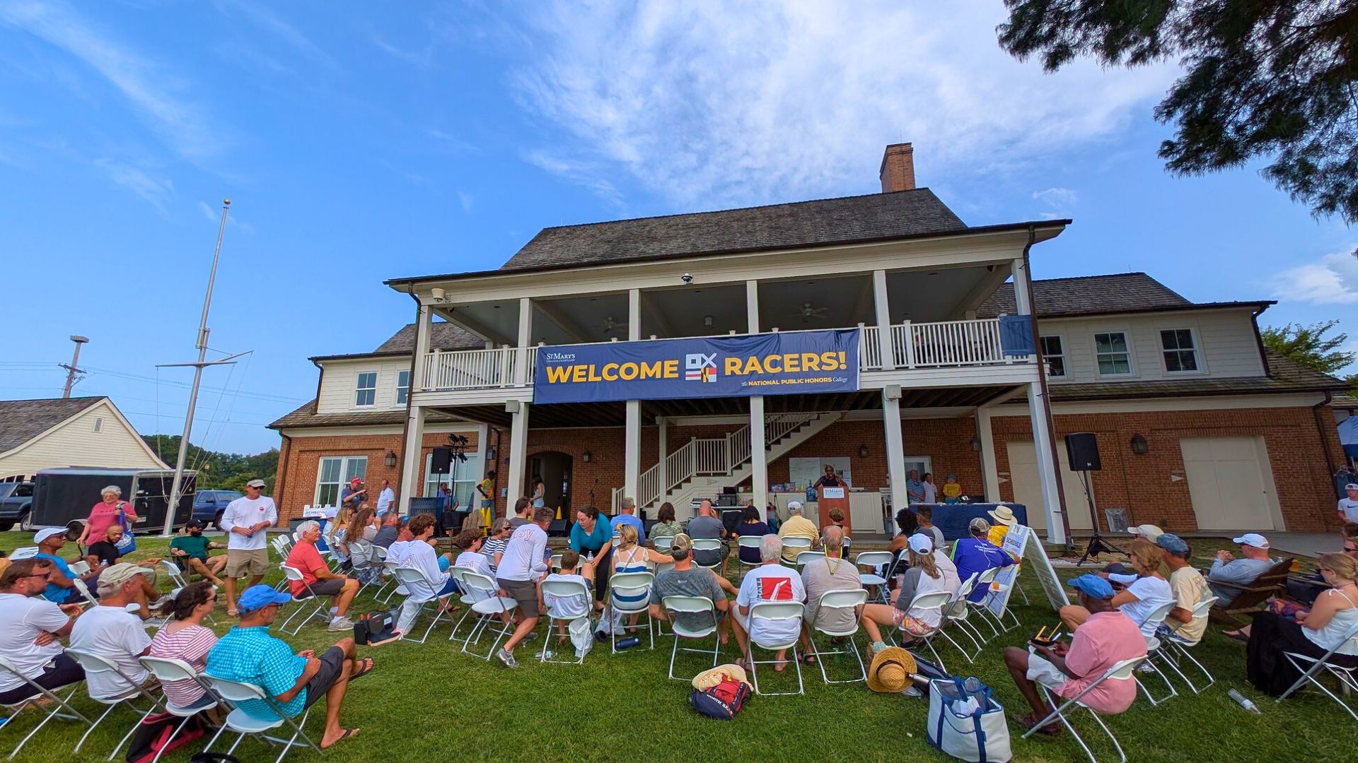 A group of people sits on white chairs facing a stage with a "WELCOME RACERS!" banner outside a two-story brick building on a sunny day.