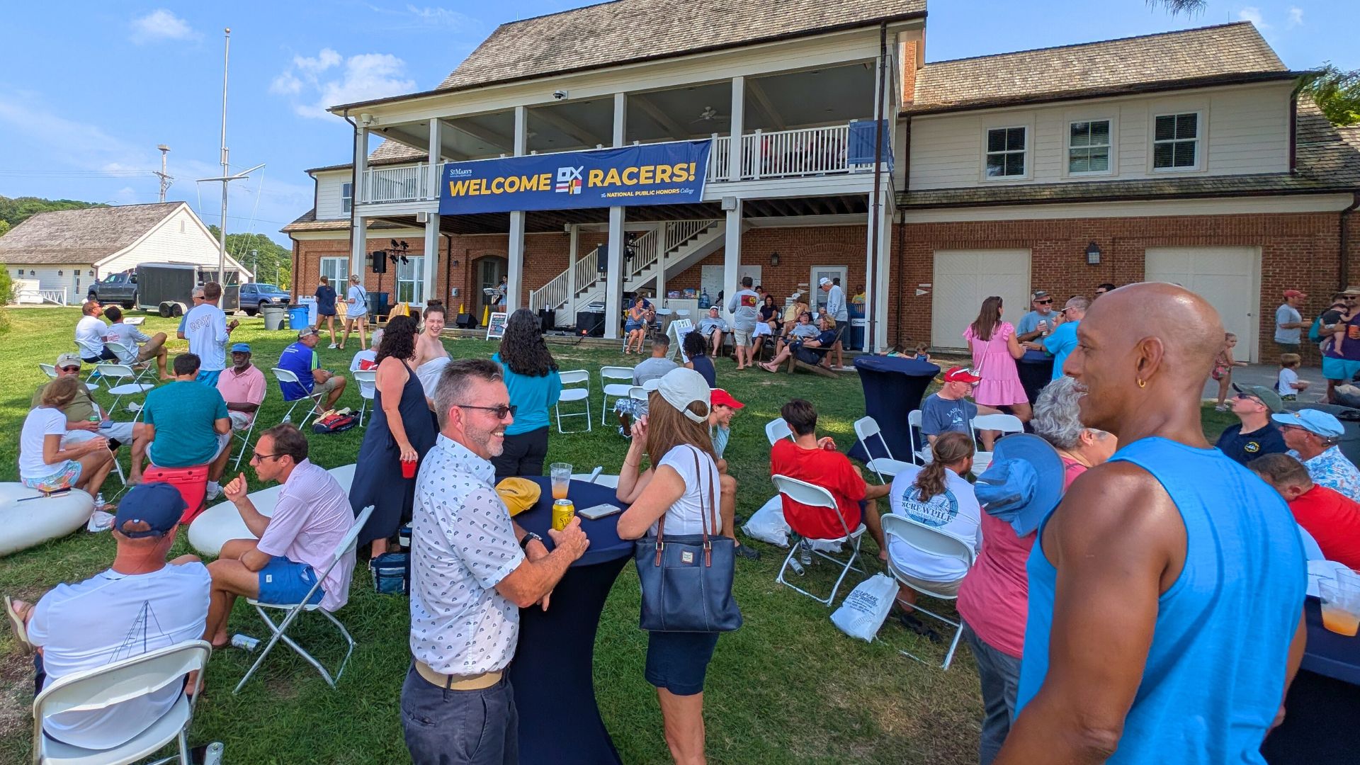 A crowd of people gathers outside a clubhouse-style building with a "Welcome Racers!" banner, sitting and standing around tables and chairs on the lawn.