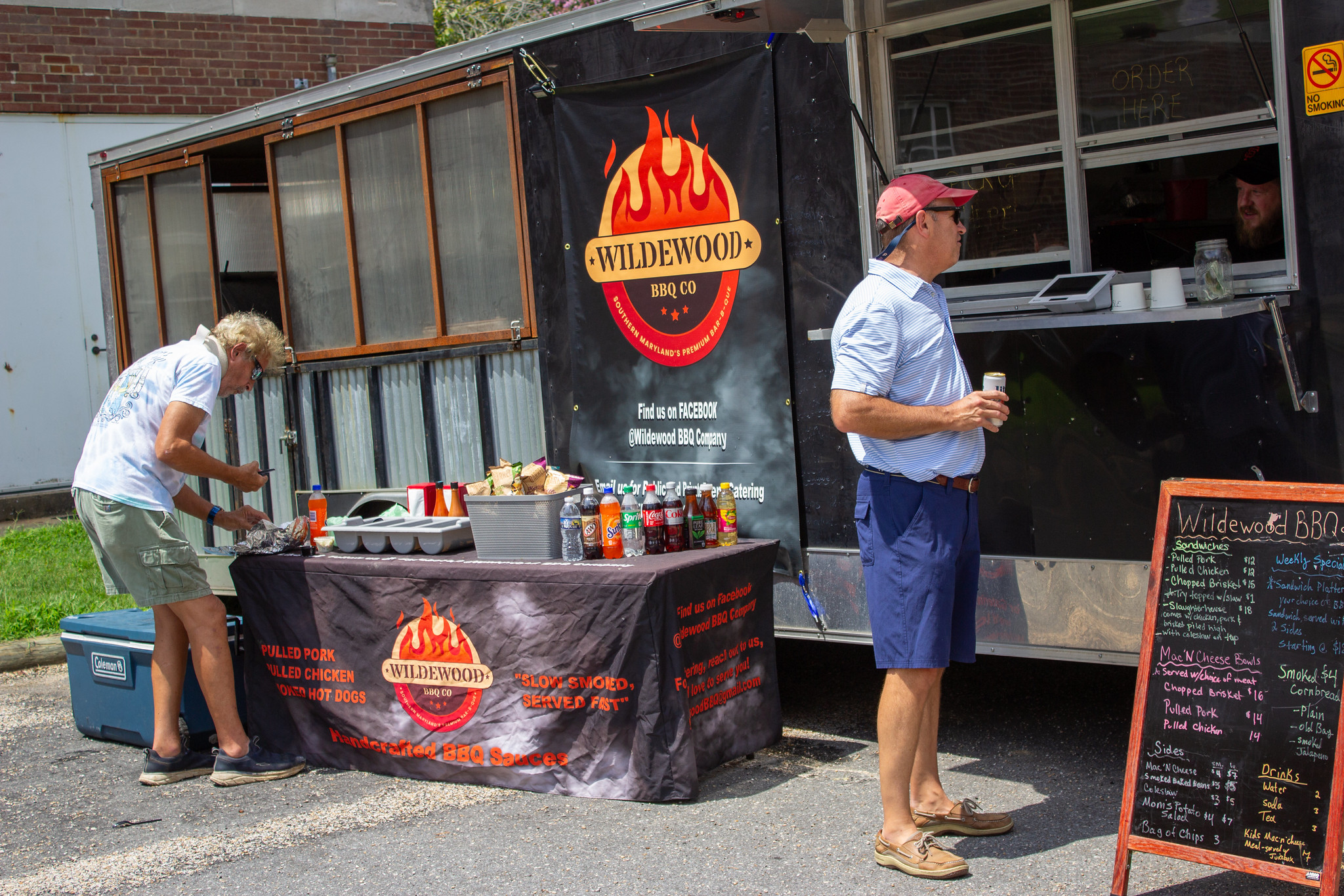 Two men stand outside a Wildewood BBQ Co. food truck; one arranges condiments on a table, while the other holds a drink near the ordering window and menu board.