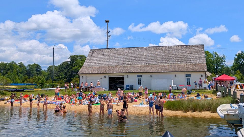 A crowd of people enjoy a sunny day at a sandy lakeside beach in front of a large white building with a wooden roof. SMCM Waterfront William E. Seale Rowing Center
