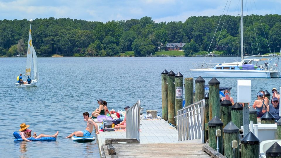 People relax on a dock and float in the water by a lake, with sailboats and trees in the background under a partly cloudy sky. SMCM Teddy Turner Waterfront