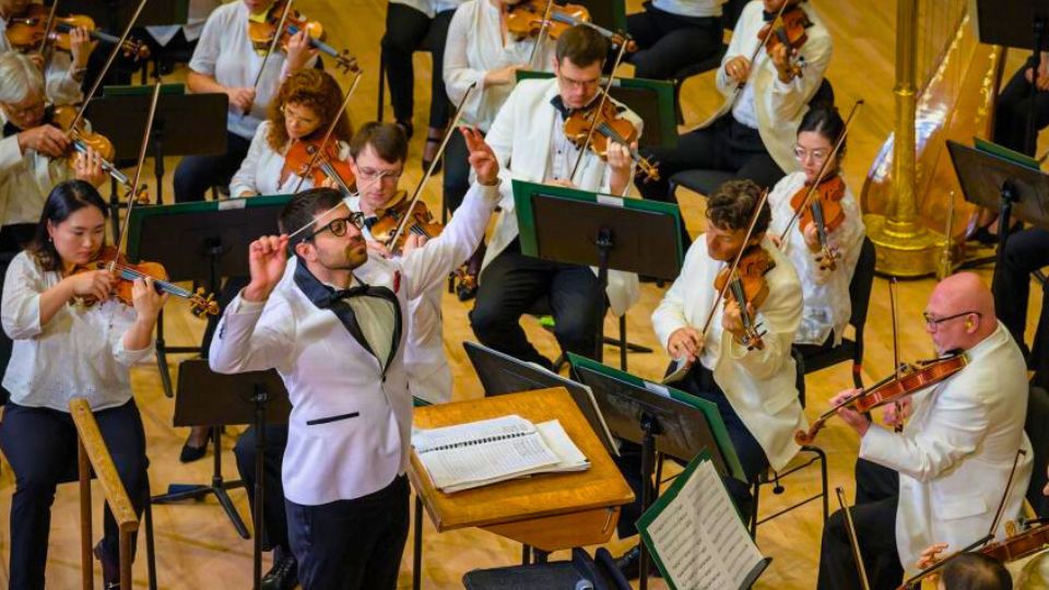 A conductor leads an orchestra of violinists and musicians dressed in white jackets, performing on a wooden stage with sheet music stands.