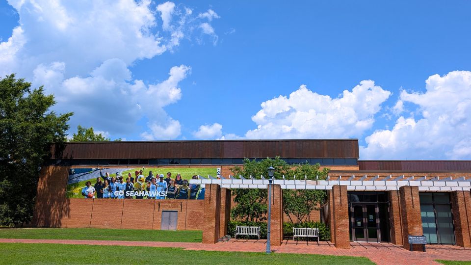 A brick building with a large banner reading "GO SEAHAWKS" on the wall, surrounded by green trees, under a blue sky with scattered clouds. MPOARC athletics and recreation facility at SMCM
