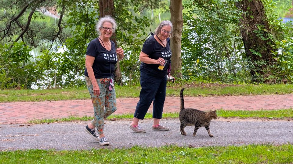 Two SMCM 1970s Alumni follow Meatball the tabby cat along a campus path, surrounded by trees and greenery.