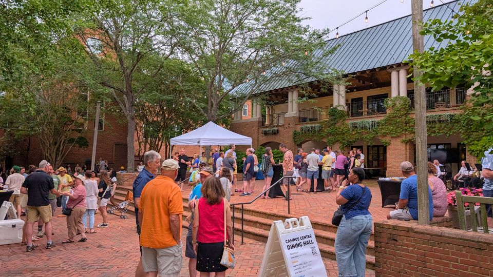 People gather outdoors in a brick courtyard with trees, a white tent, and a building with balconies; a sign reads “Campus Central Dining Hall.” SMCM Campus Center Patio