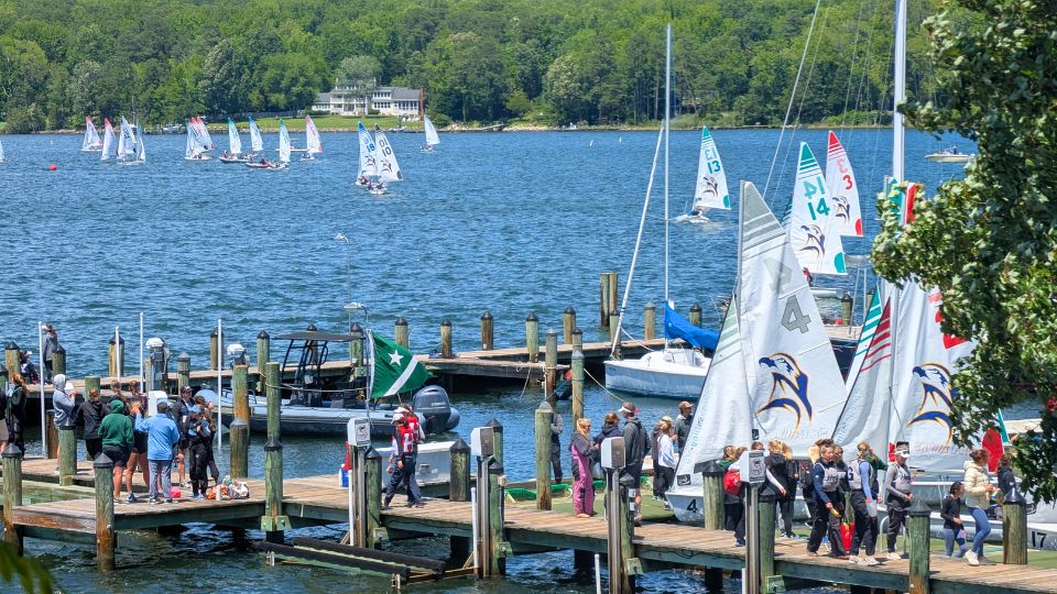 Groups of people stand on a dock by a lake filled with small sailboats; trees and a house are visible in the background. SMCM Teddy Turner Waterfront