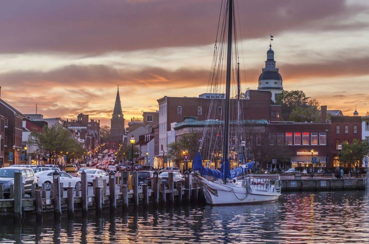 A sailboat is docked at a waterfront with historic brick buildings and a large domed structure in the background at sunset.