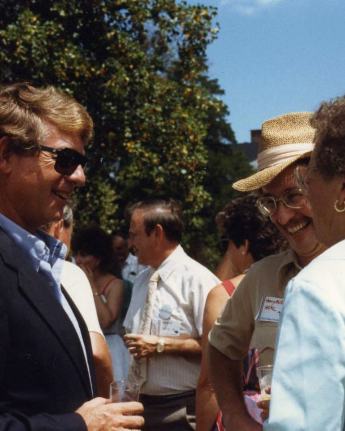 Ted Koppel and Henry Miller at the Governor's Cup party, 1987 A group of people socializing outdoors on a sunny day; two men and a woman in the foreground smiling and talking, with drinks in hand. Ted Koppel and Henry Miller at the Governor's Cup party, 1987
