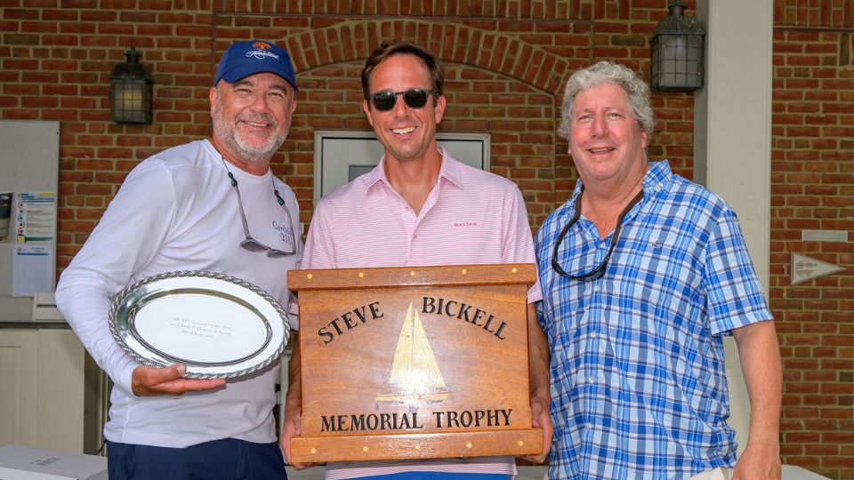 Three men standing and smiling; one holds a silver plate award, another holds a large wooden plaque reading "Steve Bickell Memorial Trophy." Brick wall and door in the background.