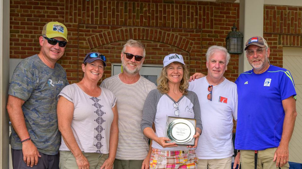 Six adults stand together smiling; the woman in the center holds a silver plate award. Brick wall and door frame are visible in the background.
