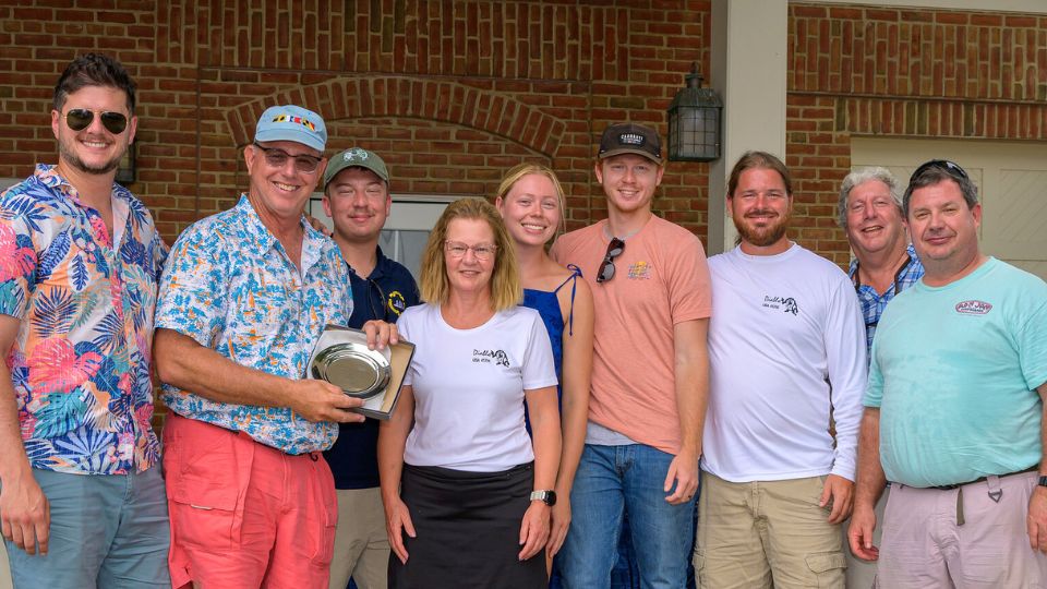 A group of nine adults stand together outside, smiling. One man holds a metal plate or trophy. Most people wear casual summer clothing. Brick and white building in the background.