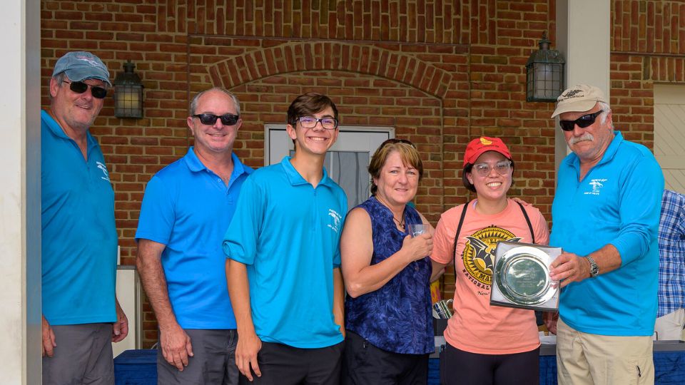 Six people stand together posing for a photo; two women in the center hold a silver tray award. Most group members wear blue shirts, with a brick wall in the background.