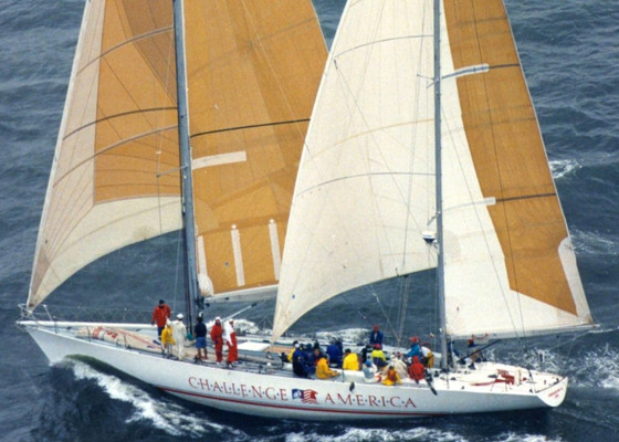 A large sailboat with tan sails labeled "Challenge America" sails on the ocean, crew members in colorful jackets are visible on deck.