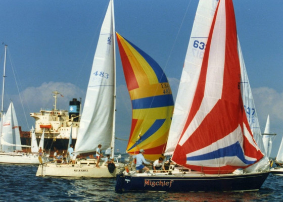 Several sailboats with colorful sails race on the water, with a large cargo ship and more boats visible in the background. One is named Mischief