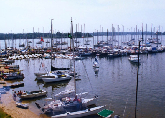 Dozens of sailboats and motorboats are docked at a marina on a calm lake, with people visible on some boats and along the shore. Governor's Cup 1991