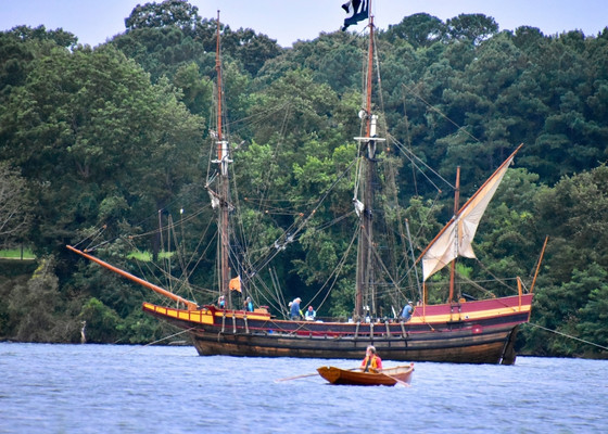 A wooden sailing ship with multiple masts, The Maryland Dove, is anchored near the shore by Church Point as a small rowboat with one person passes in the foreground. Trees line the background.