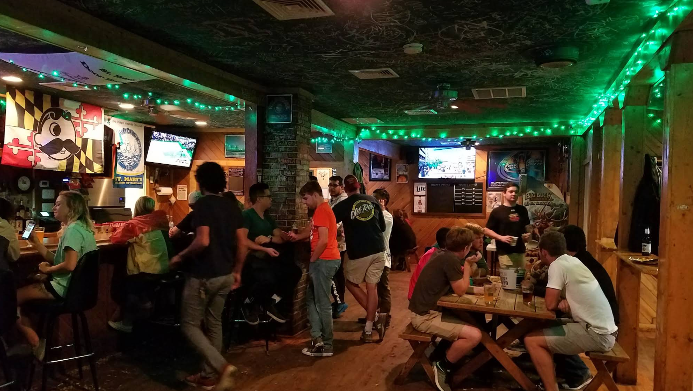 A group of people socialize at a dimly lit bar decorated with green string lights, with several TVs showing sports in the background.
