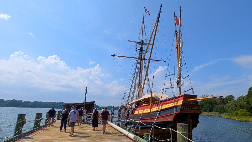 A group of people walk along a wooden dock toward a historic sailing ship, The Maryland Dove, moored by a lake under a bright blue sky at Historic St. Mary's City