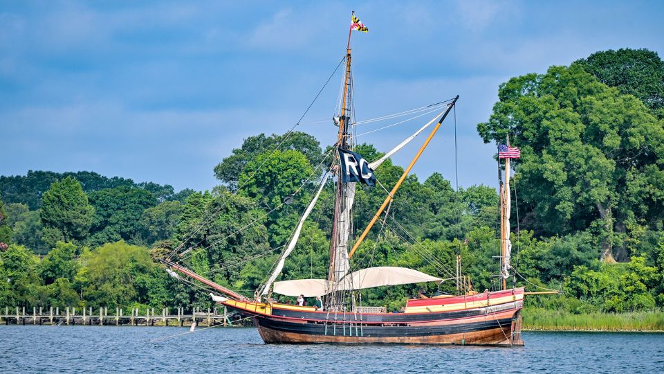 The Maryland Dove, a historic wooden sailing ship with tall masts and flags is anchored near a wooded shoreline on a calm, sunny day, acting as RC ship during the SMCM Governor's Cup Yacht Race