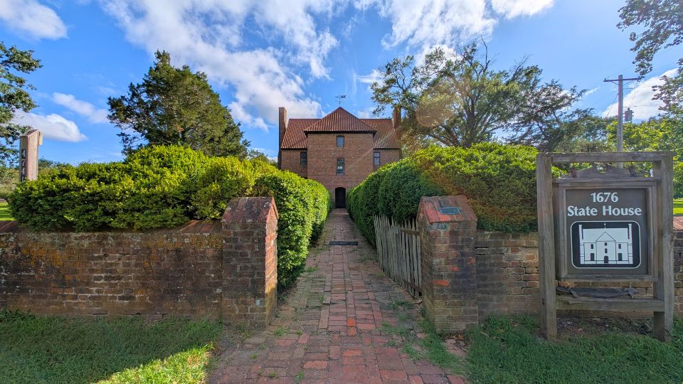 A brick pathway leads to the entrance of a historic brick building labeled "1676 State House," surrounded by trimmed hedges and a wooden sign with the building's name and illustration, at Historic St. Mary's City, MD.