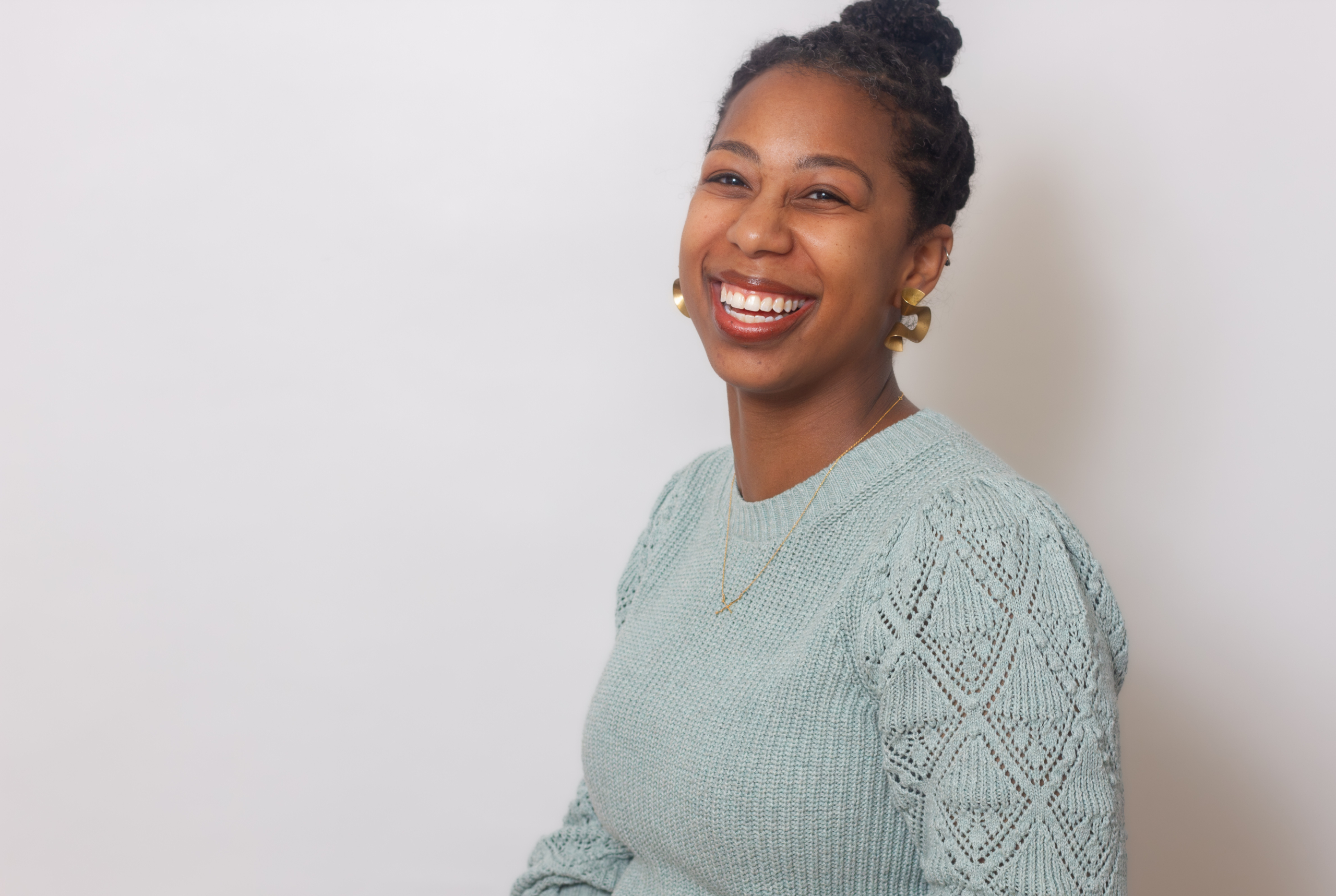 A woman with braided hair in a bun, wearing a light blue knit sweater and gold earrings, smiles at the camera against a plain white background.
