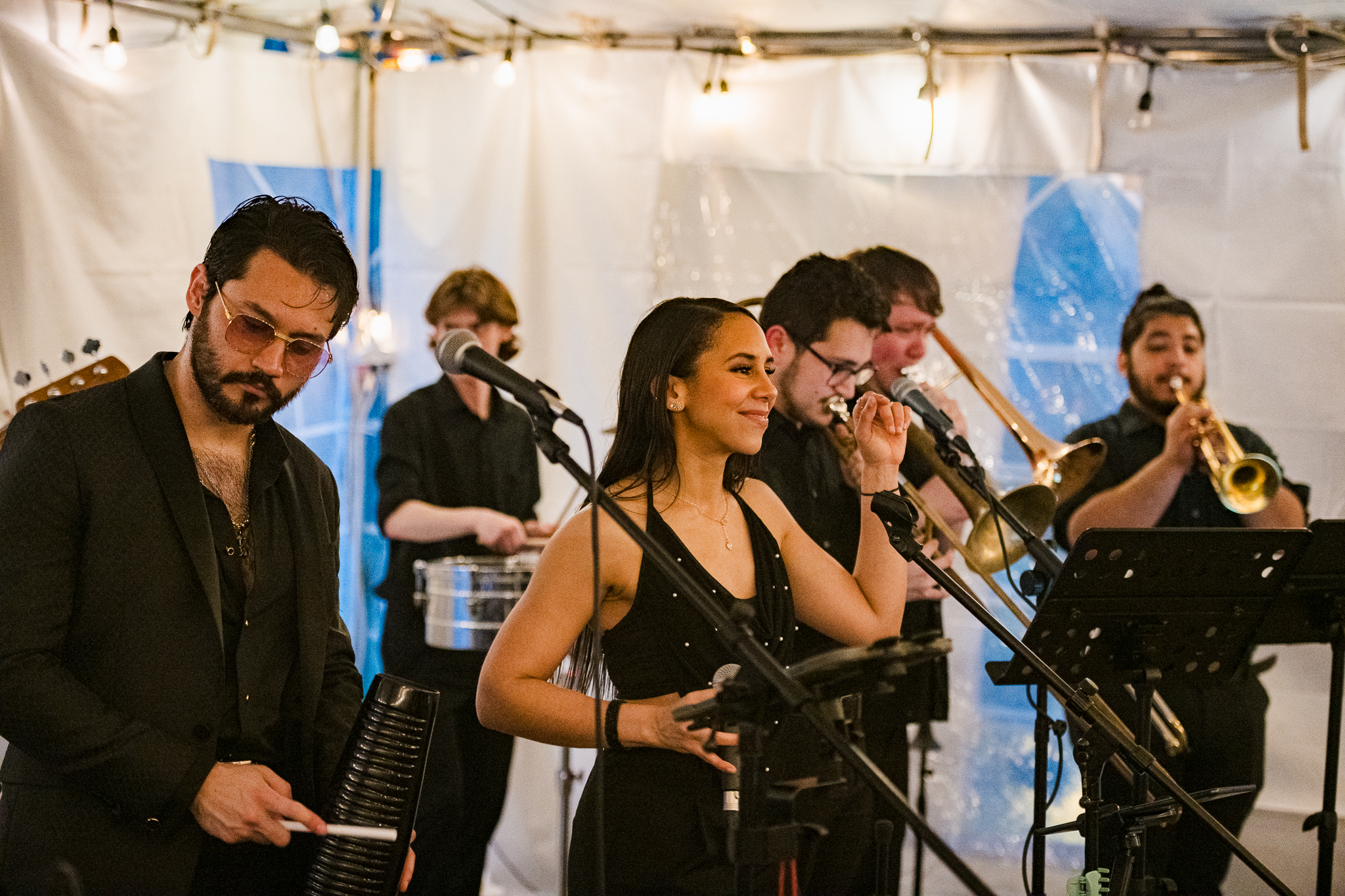 A band performs indoors with multiple musicians playing percussion, trombone, trumpet, and singing into microphones under string lights.