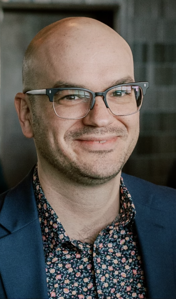 A man with glasses, and light facial hair is wearing a blue blazer and a floral shirt, smiling indoors.