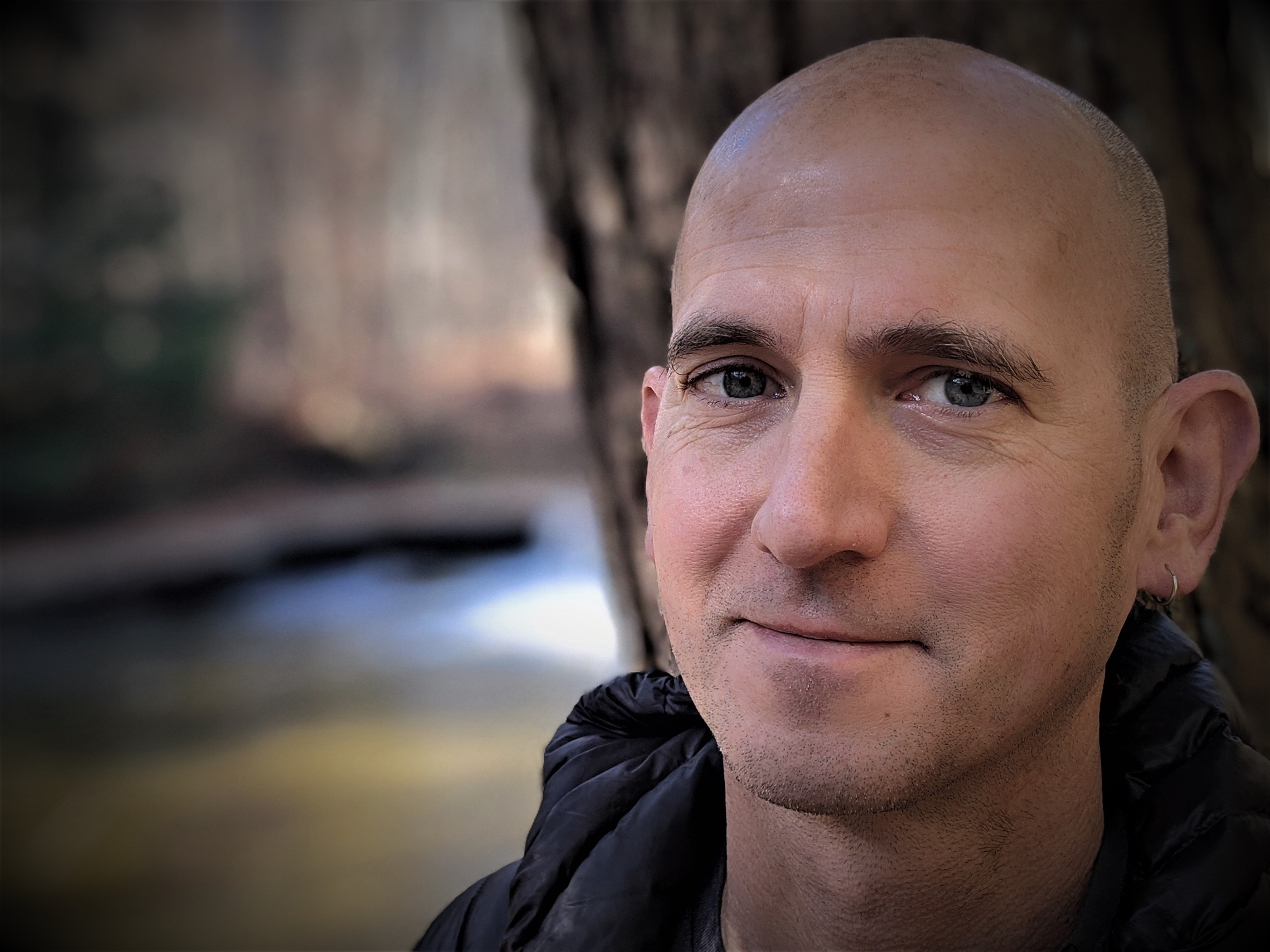 A person standing outdoors with a neutral expression, wearing a dark jacket. Behind them, a scenic view of a lake surrounded by autumn foliage and trees under a cloudy sky.