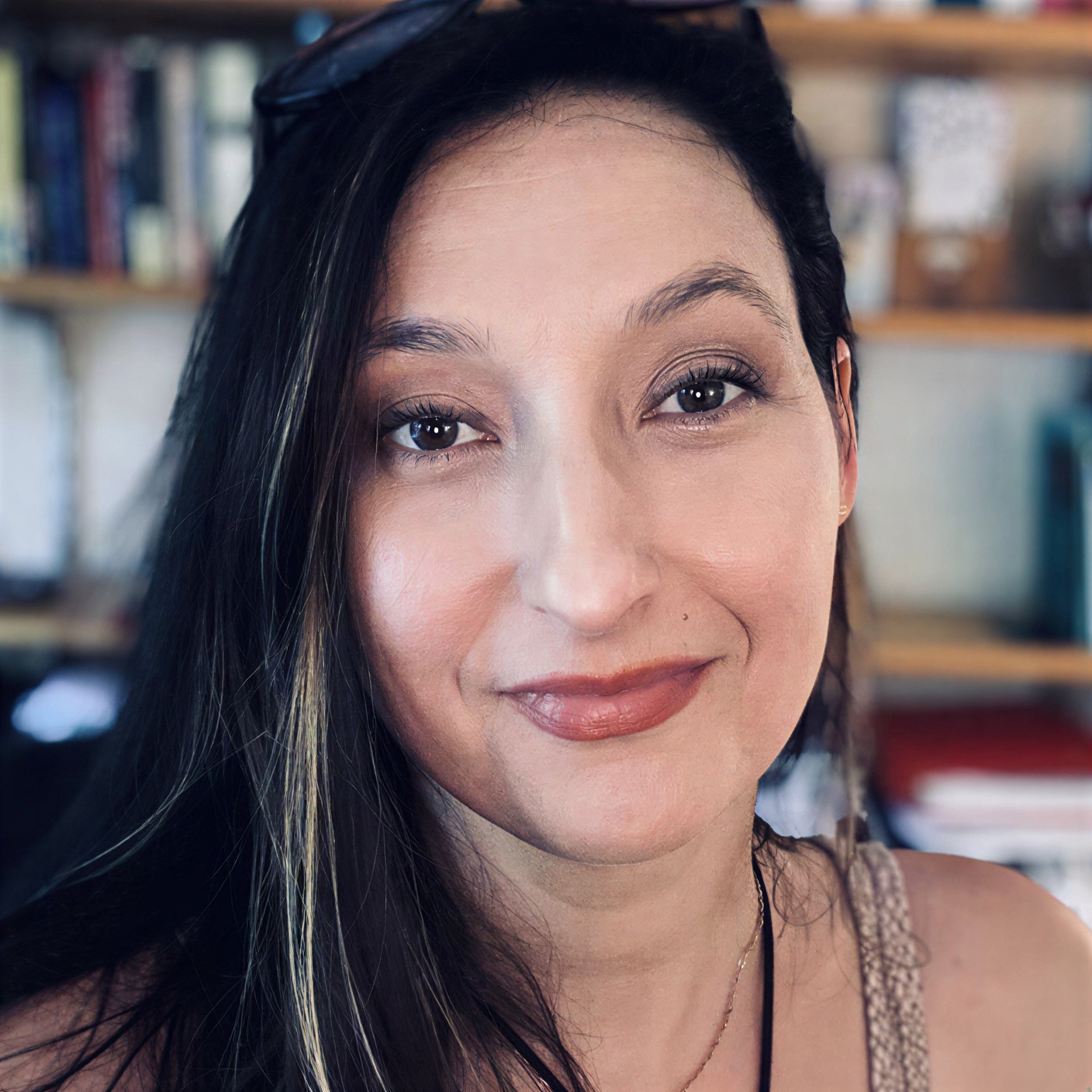 A woman with long brown hair and light makeup is smiling softly at the camera, with bookshelves in the blurred background.
